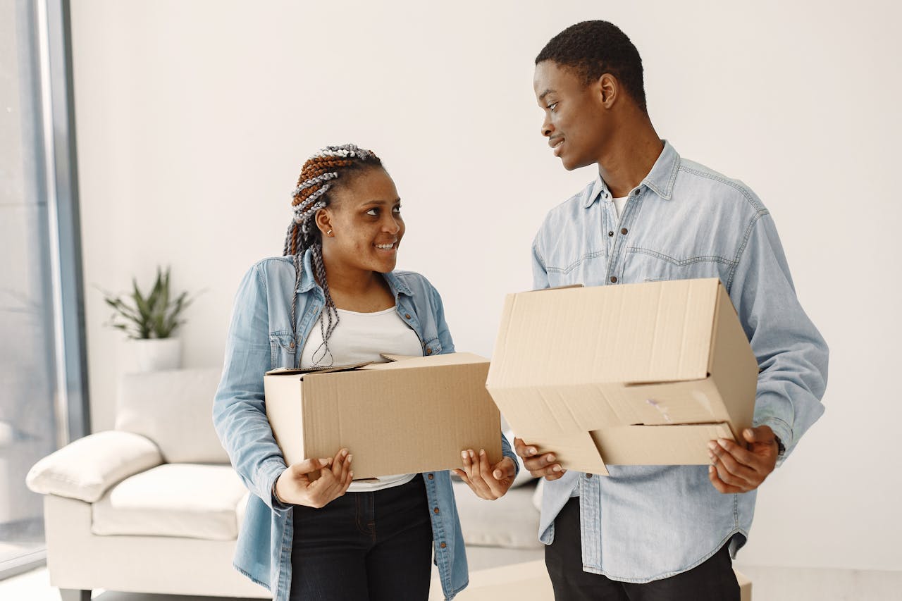 A young couple moving in, smiling while holding boxes in a bright living room.