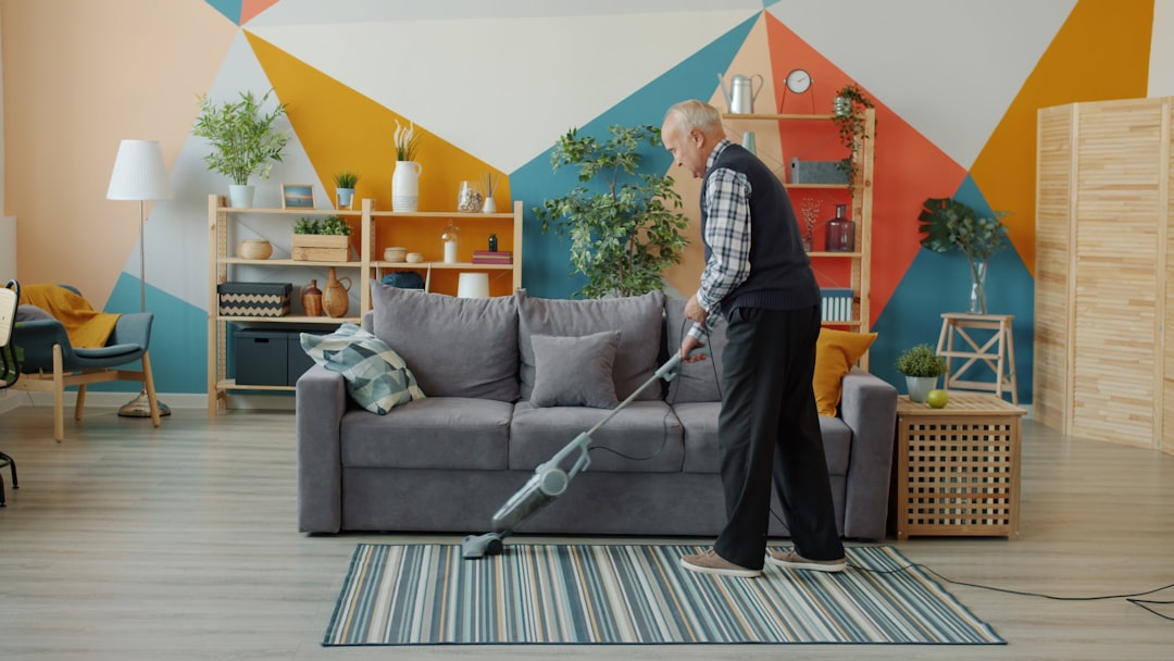 Senior man vacuuming carpet at home with vacuum cleaner doing housework alone concentrated on clean-up activity. People, housing and household concept.